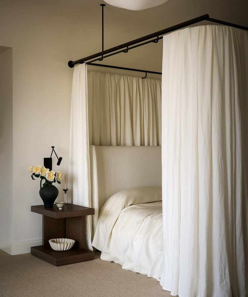 neutral minimalist bedroom with a canopy bed and white bedding, and a dark wooden nightstand