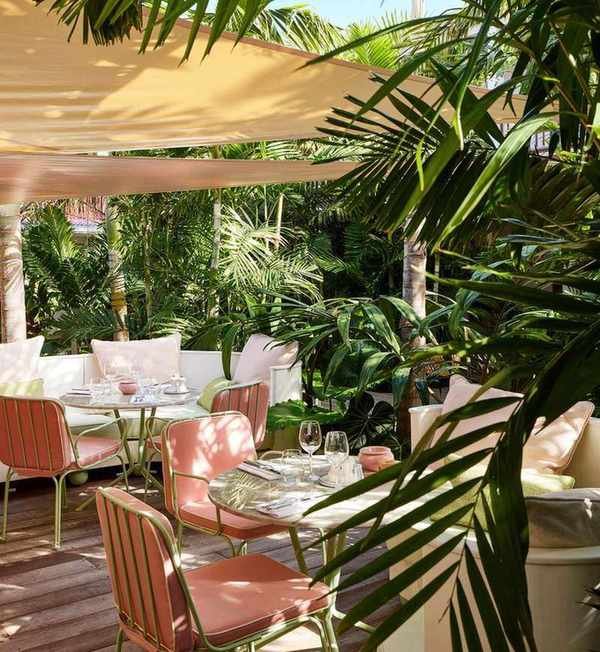 Two outdoor dining tables surrounded by greenery with pink chairs and a tan shade hanging above