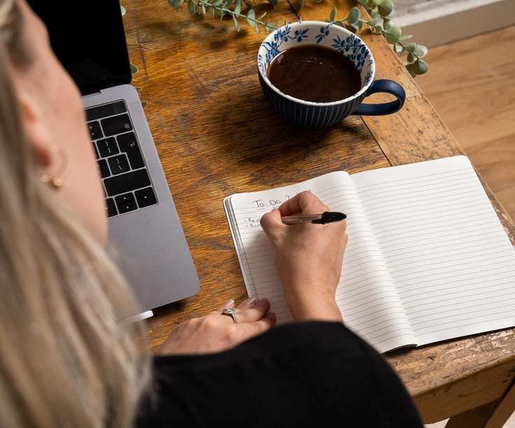 Writing a to do list in a notepad next to a laptop
