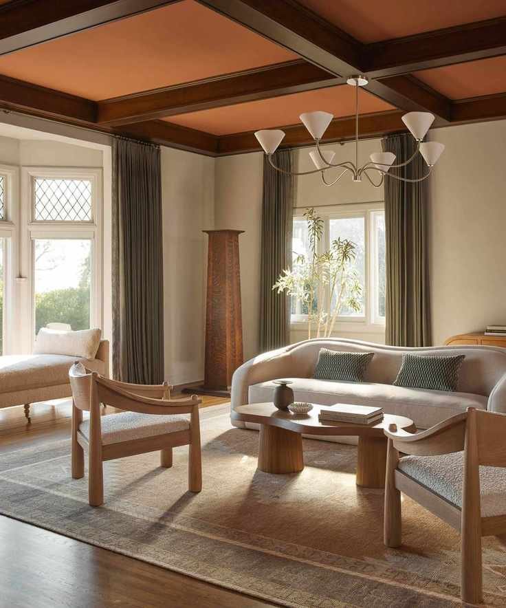 neutral living room with curved boucle sofa, oak coffee table, accent chairs, a large silver chandelier and a beamed wooden ceiling painted orange