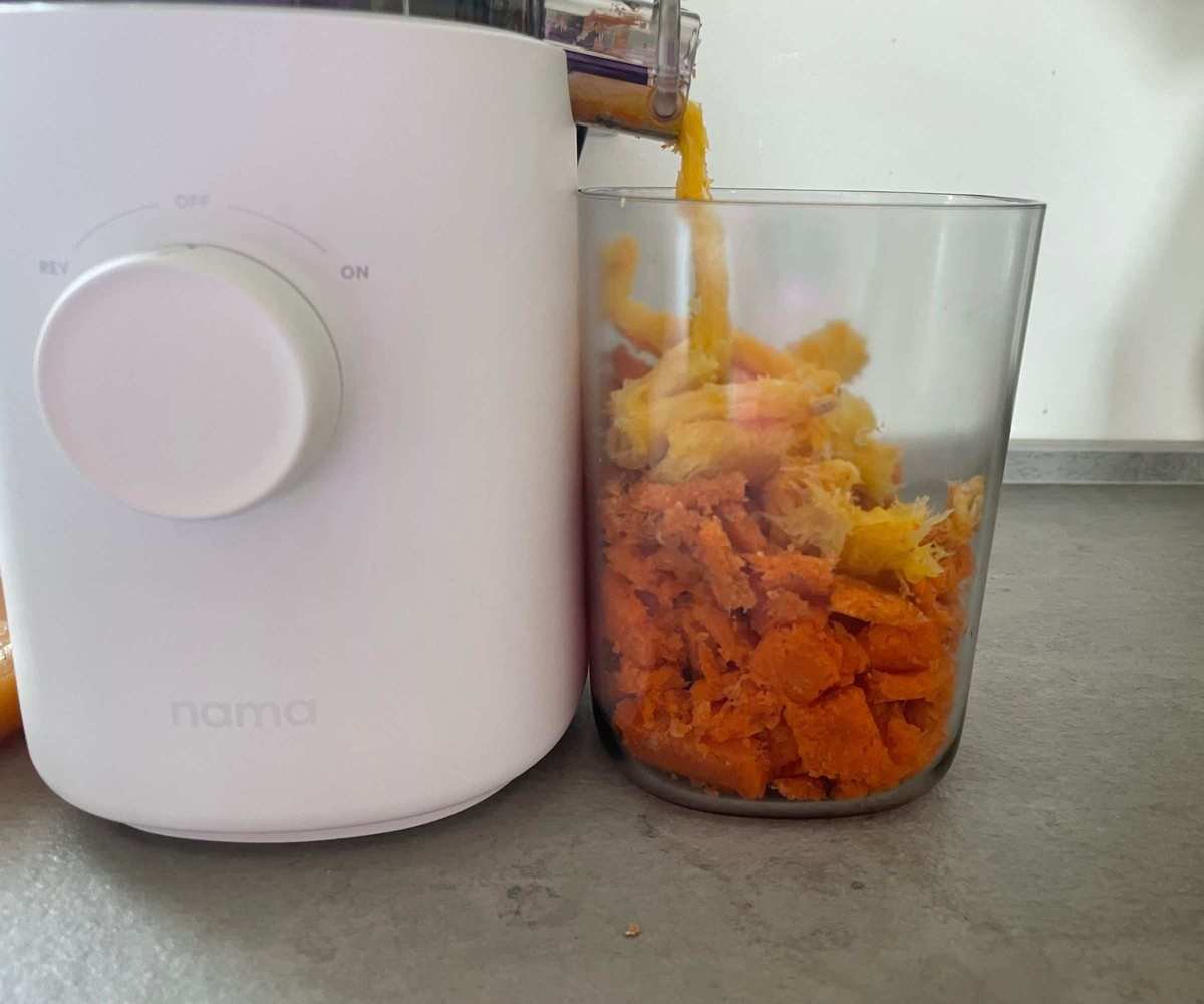 Yellow pulp being funnelled on top of orange pulp into a clear plastic beaker next to the white base of a juicer on a grey counter.
