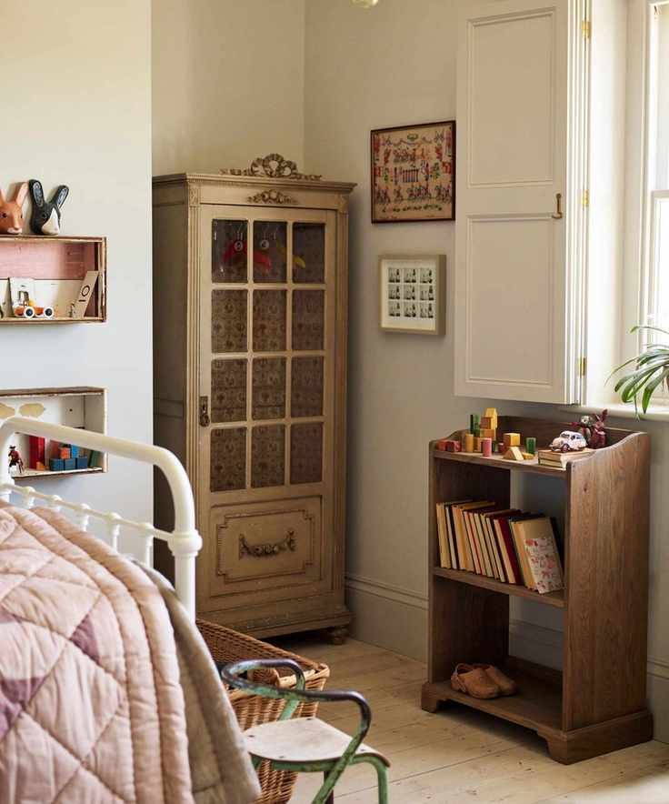 A child's bedroom with a small wooden closet with glass doors, and a wooden bookshelf with books and toys stacked on top.