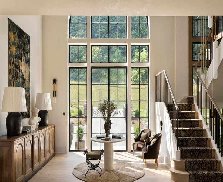 Large open cream entryway with a wooden console table on the left hand wall. On it is two black lamps with white shades and above is a vintage tapestry. In the middle is a circular rug with a small circular table.