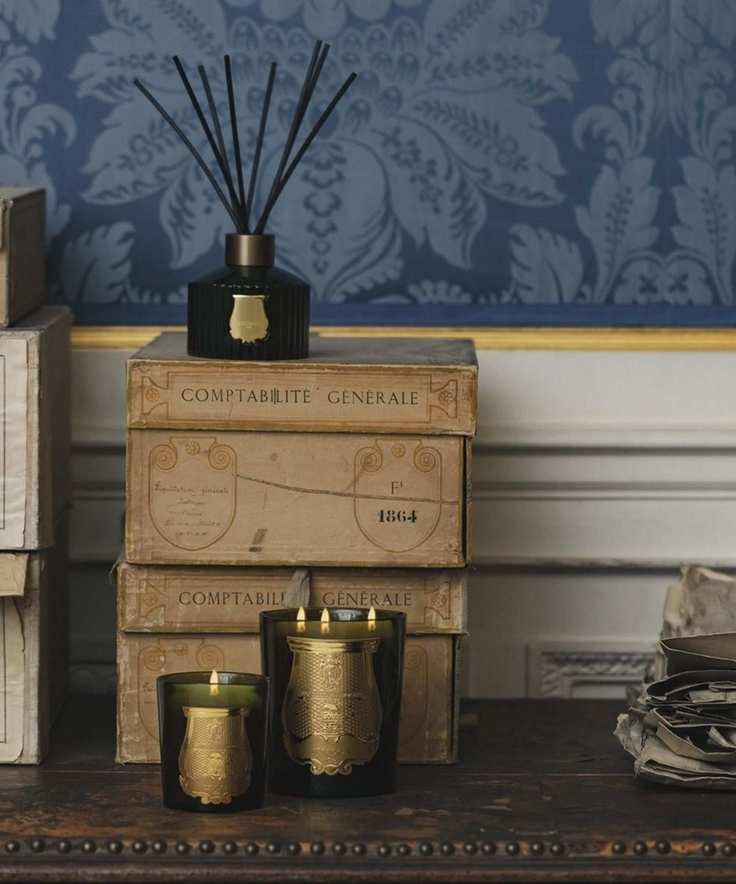 A black Trudon Ernesto diffuser with black reeds on a stack of decorative boxes on a wooden table. In front of the boxes are two lit green Trudon candles with gold labels, and behind is a white panelled wall, with a gold strip and blue patterned wallpaper above.