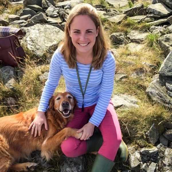 Dr Aimee Warner is a white women, with shoulder length dark blonde hair. She is pictured outdoors, smiling, sat on mossy rocks with a very happy dog which is resting its paw on her leg.