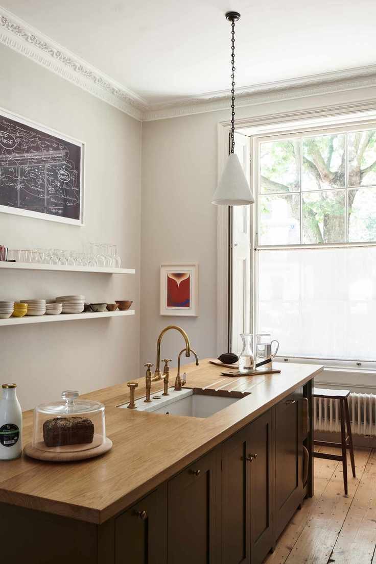 kitchen with wooden topped island with sink, open plan shelving on one wall, window, stool, wooden floor, pendant light, artwork