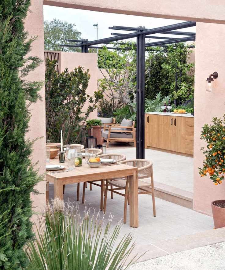 Outdoor dining area surrounded by pink walls and greenery, on concrete floor with black pergola and kitchen area in background.