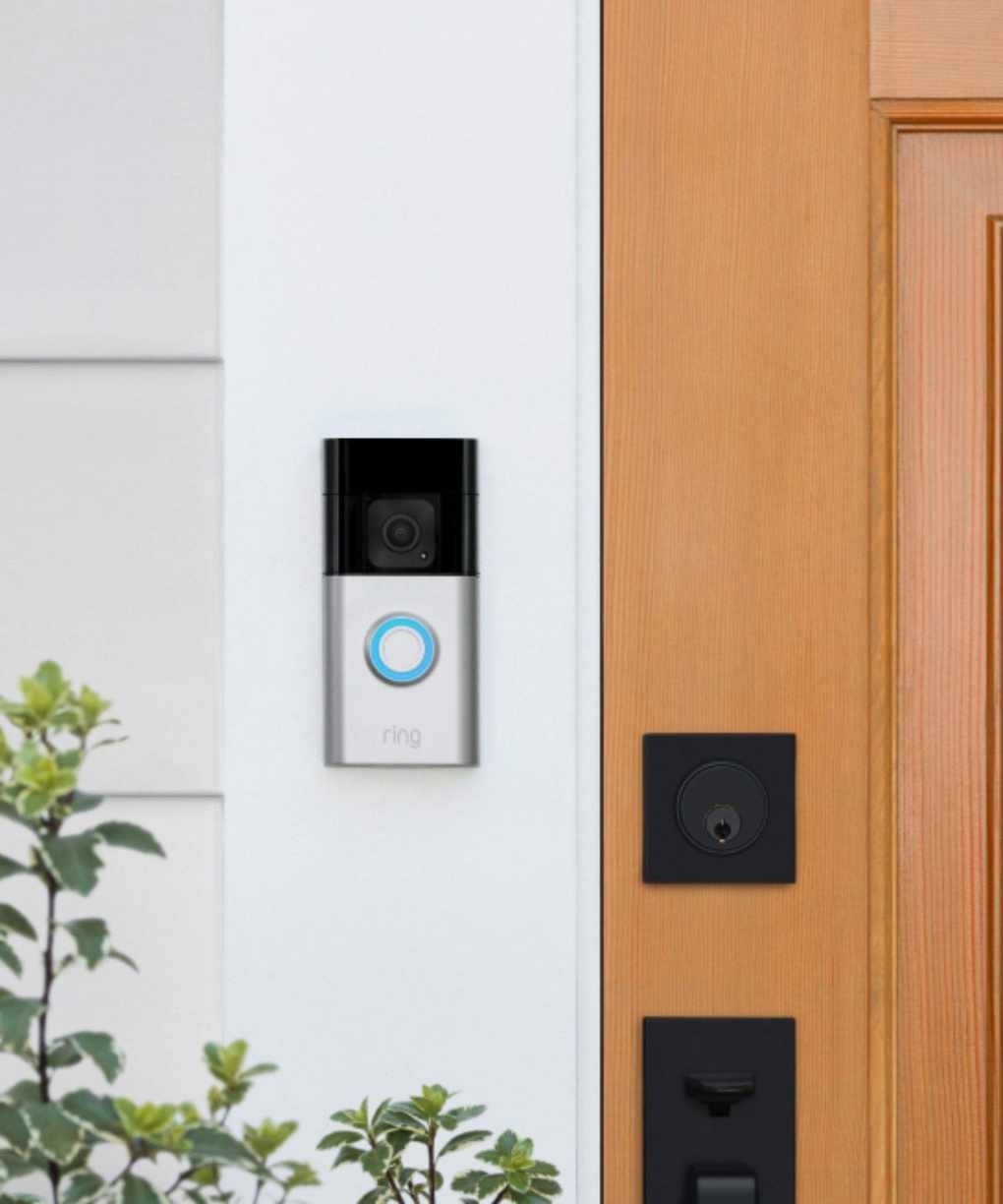A black and silver Ring Battery Doorbell Plus mounted on a white wall next to a wooden front door, with leaves in the foreground