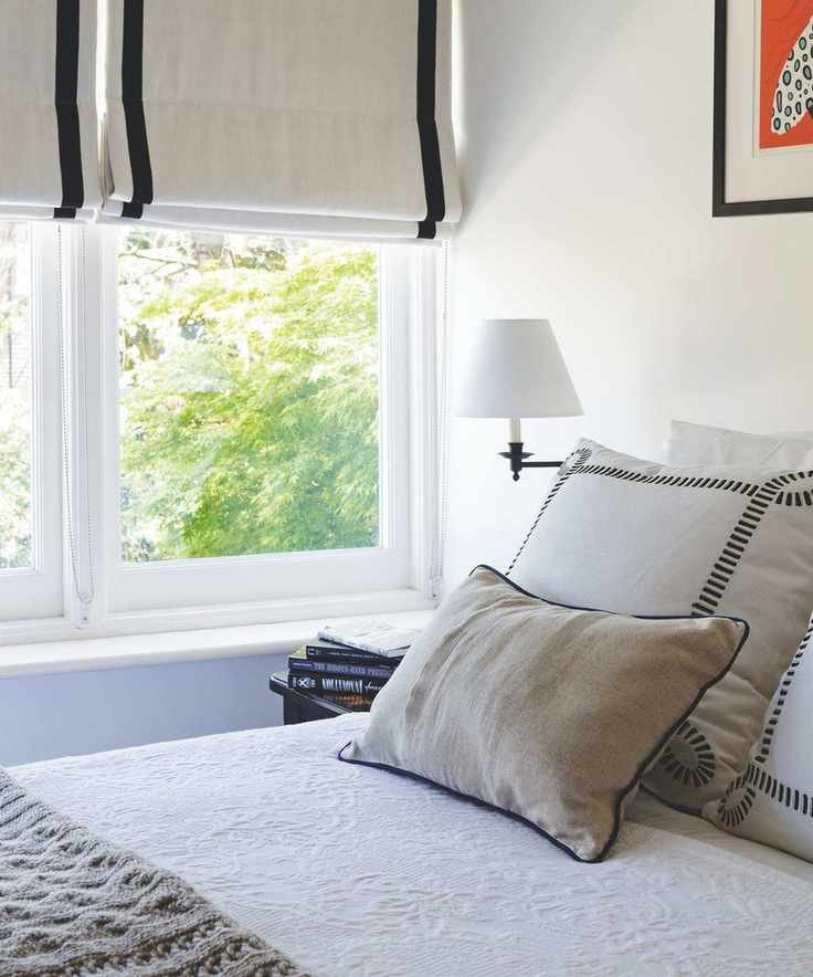 A white bedroom with a bed with white bedding in the foreground, with a grey cushion at the foot and two large square white and black cushions at the head, with a rectangular neutral beige cushion leaning up against them. To the left of the bed is a white wall lamp, and the corner of a bedside table with a stack of books on it is seen. Behind is a large window with green leaves seen beyond, and a cream and black blind.