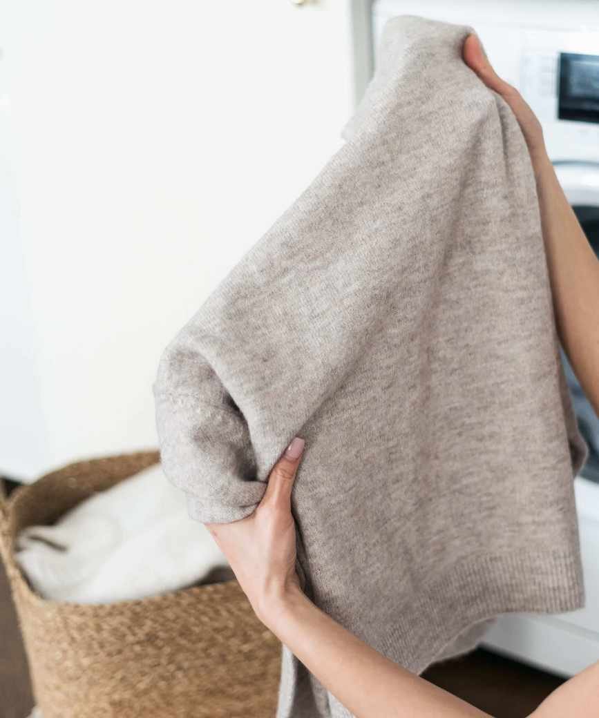 Someone holding up a gray wool sweater in front of a white washing machine and laundry basket