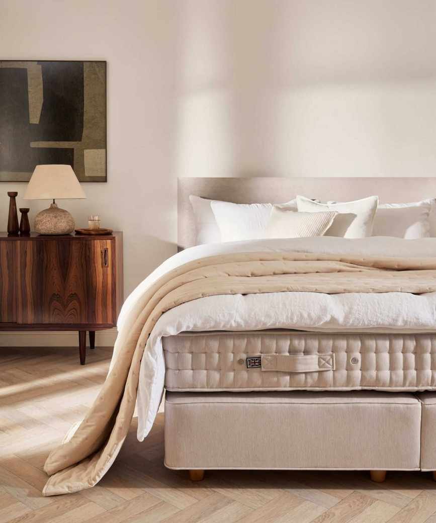neutral bedroom with an unmade bed showing the mattress, duvet, and pillows with a linen bed frame and mid century room decor