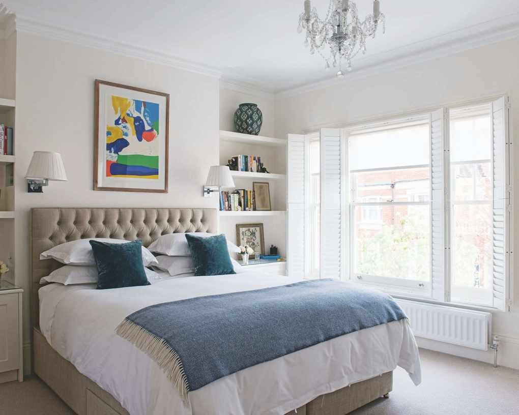 A large double bedroom with a buttoned headboard bed with white bedding, a blue blanket and blue cushions, white walls with colorful framed artwork above the bed, shelves with miscellaneous items such as books, framed pictures and vases, and large windows with shutters.