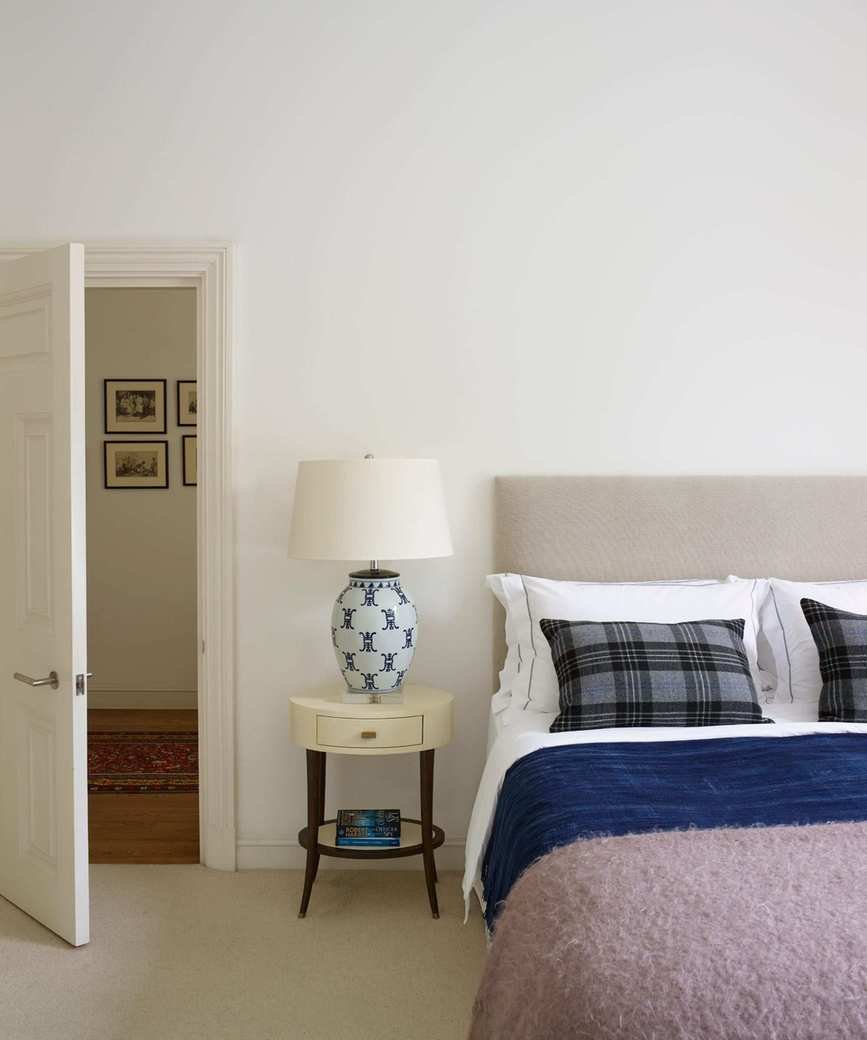 White master bedroom with double bed covered in pillows, blankets and throws, and bedside table with lamp and open door to the landing in a redecorated period house.