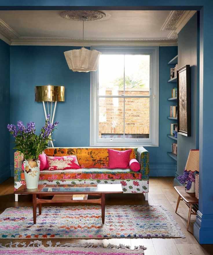 Mid blue living room with bright window, alcove shelving, brightly colored fabric sofa, multi colored rugs, mid century coffee table and vase of purple flowers