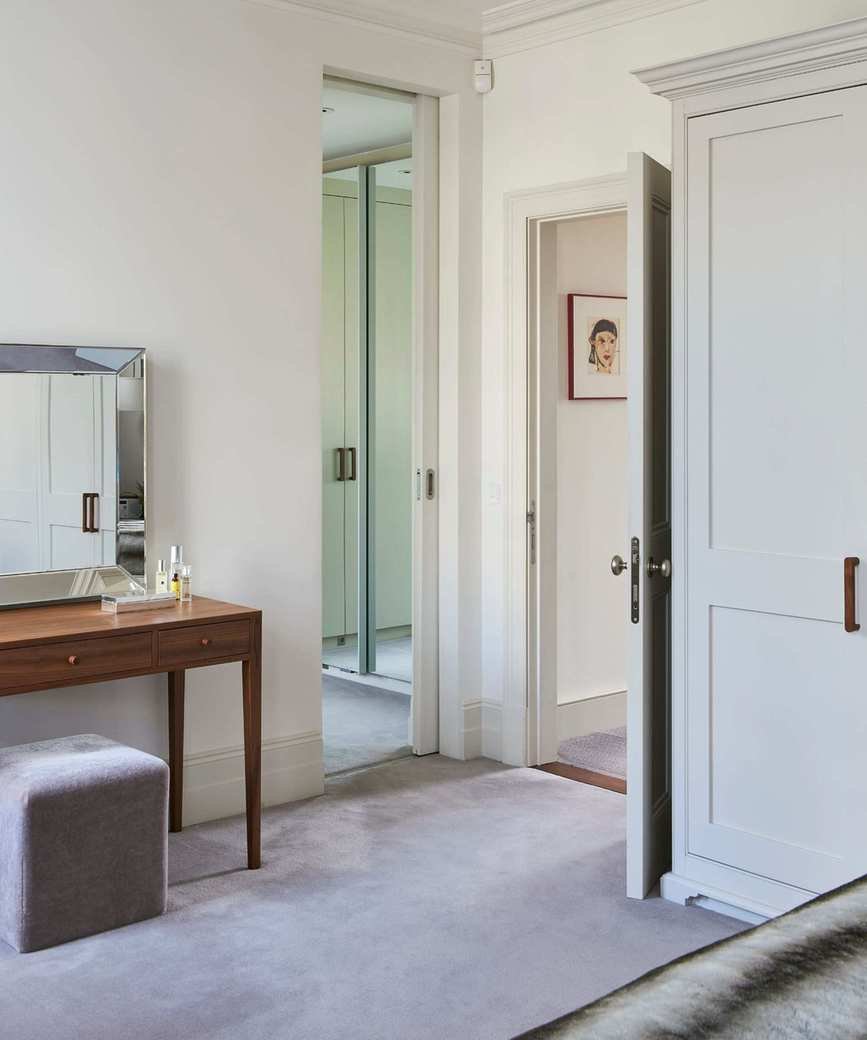 A white bedroom with a built-in closet, grey carpet and brown dressing table with a large mirror and grey velvet stool.