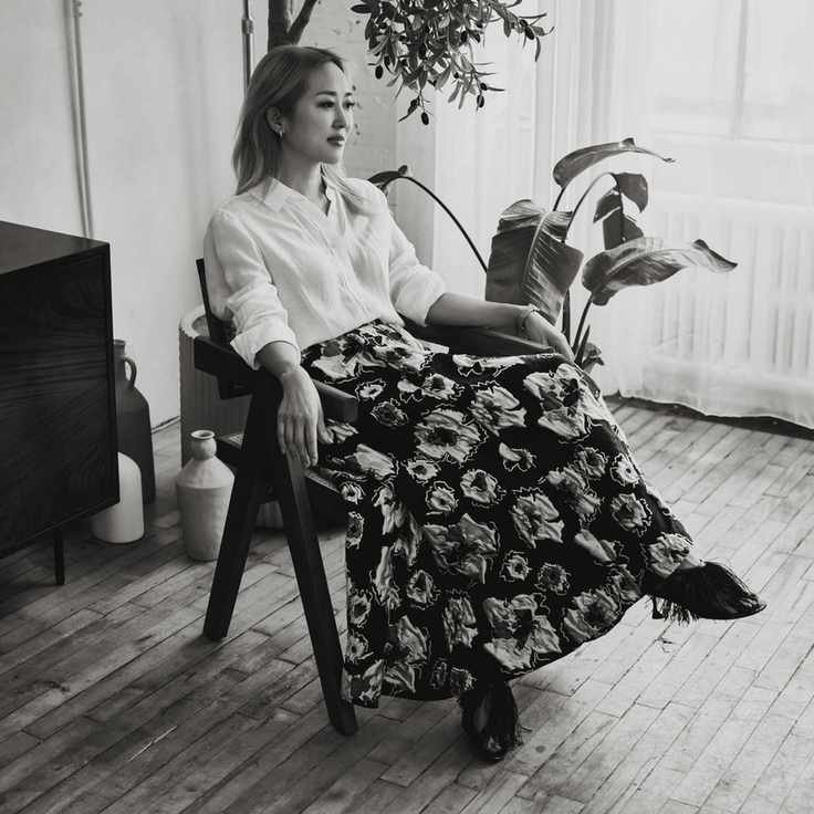 A black and white image of Maiko Shimazaki wearing a white skirt and black and white patterned skirt, sitting in a chair.