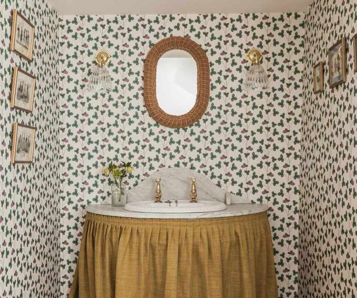Vintage sink with upholstery curtain, marble basin and vintage wallpaper with rattan mirror, glass sconces and small framed pictures
