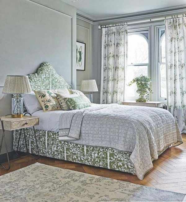 A pale green bedroom with windows, sage panelled walls, a green patterned double bed, patterned bedside tables, and patterned lamps. The floor is light wood and polished, with a large patterned rug.