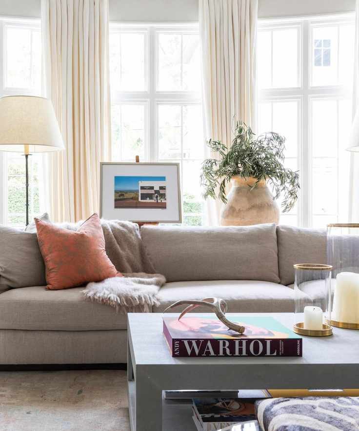 living room with cream drapes, gray couch, coffee table with books and candles, rust couch pillows, floor lamps