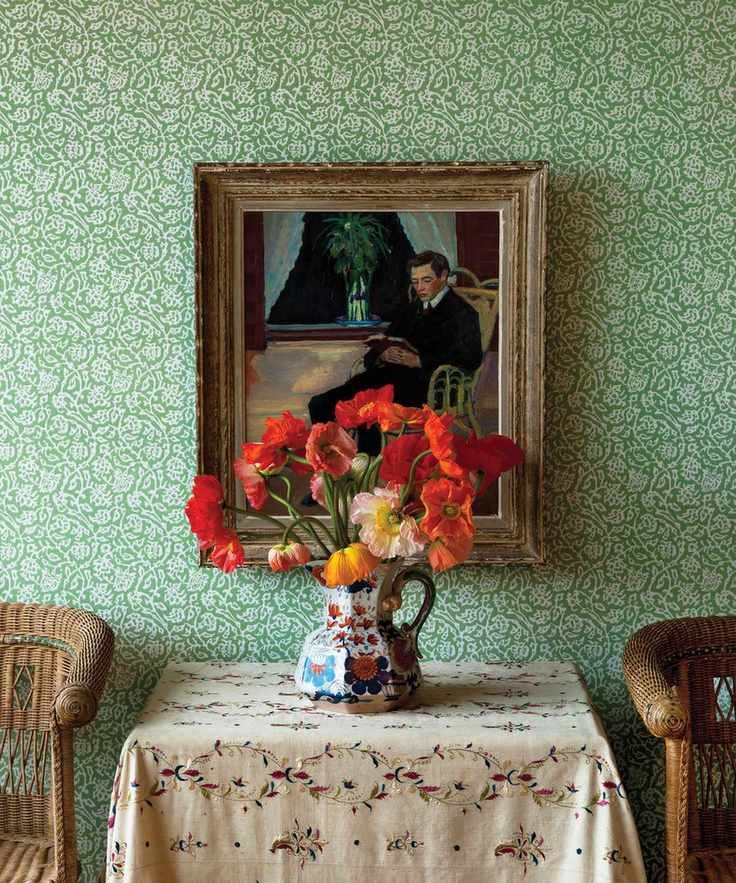 Traditional hallway with green, floral wallpaper, cream tablecloth, antique jug with red flowers, and rattan chairs