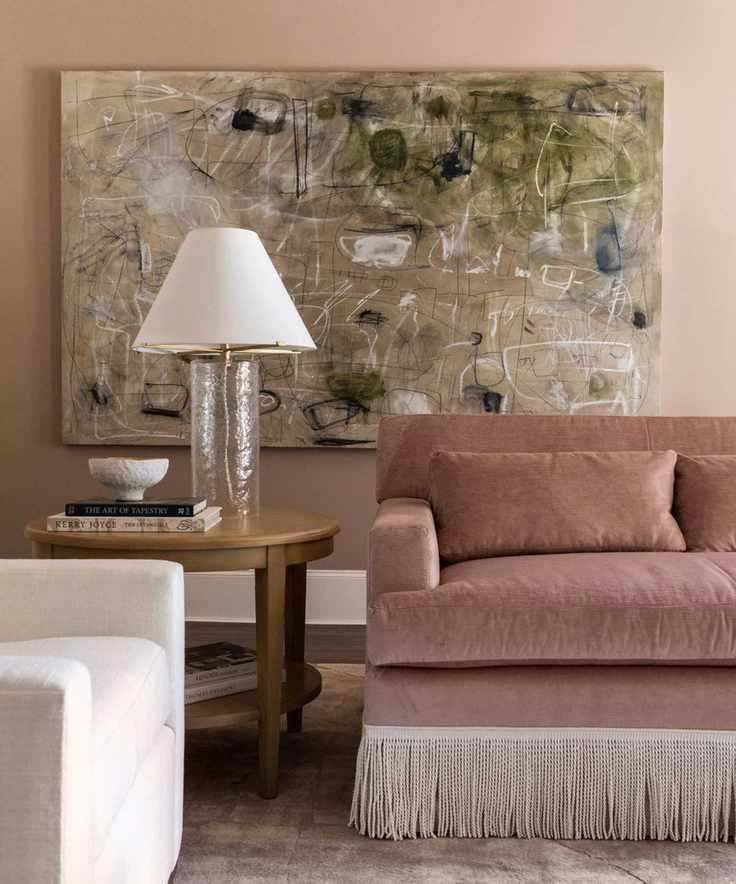 Pink living room with pink velvet fringed couch, large artwork, wooden side table and glass lamp