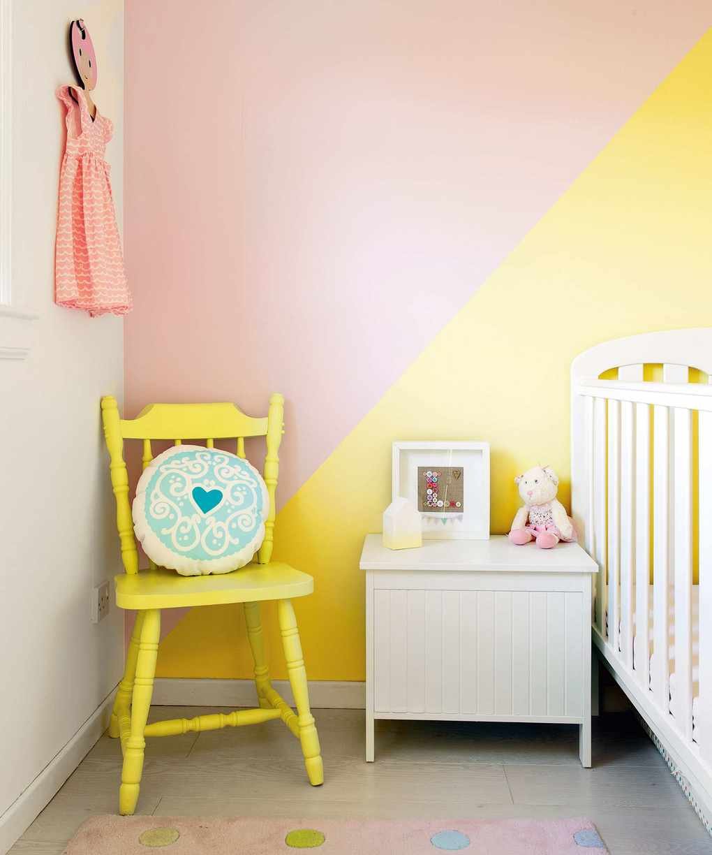 A child's nursery featuring a white cot and yellow wooden chair with a diagonal pink and yellow wall