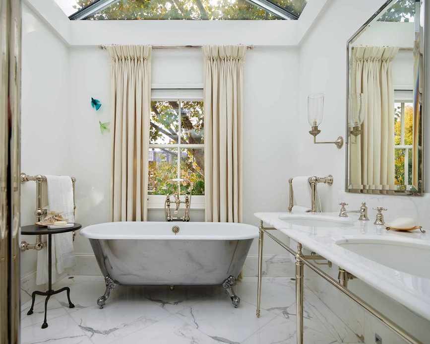 A bathroom design featuring a rolltop bath under a window, marble flooring, and a double vanity
