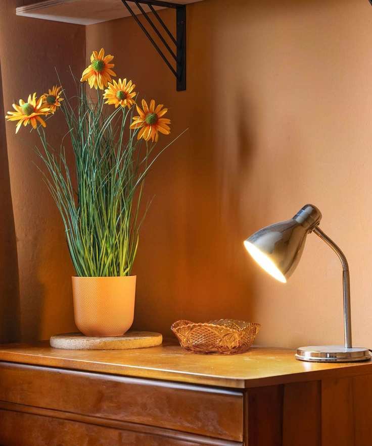 A tabletop lamp is lit on top of a chest of drawers, next to a vase of artificial sunflowers and a small item tray, against brown walls.