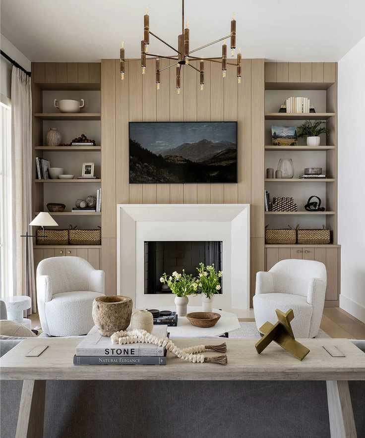 Modern living room with fireplace, tall shelving either side, tv mounted above mantel, two cream lounge chairs, floor lamp, coffee table and sofa