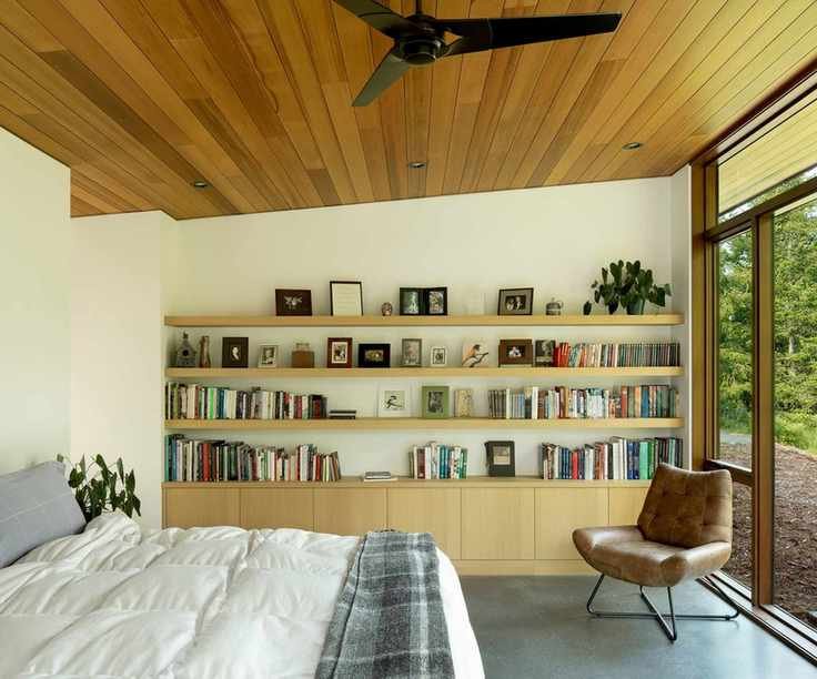 Midcentury modern bedroom with leather armchair, wooden open shelving decorated with books and pictures, and bed with white bedding, and glass doors onlooking the garden