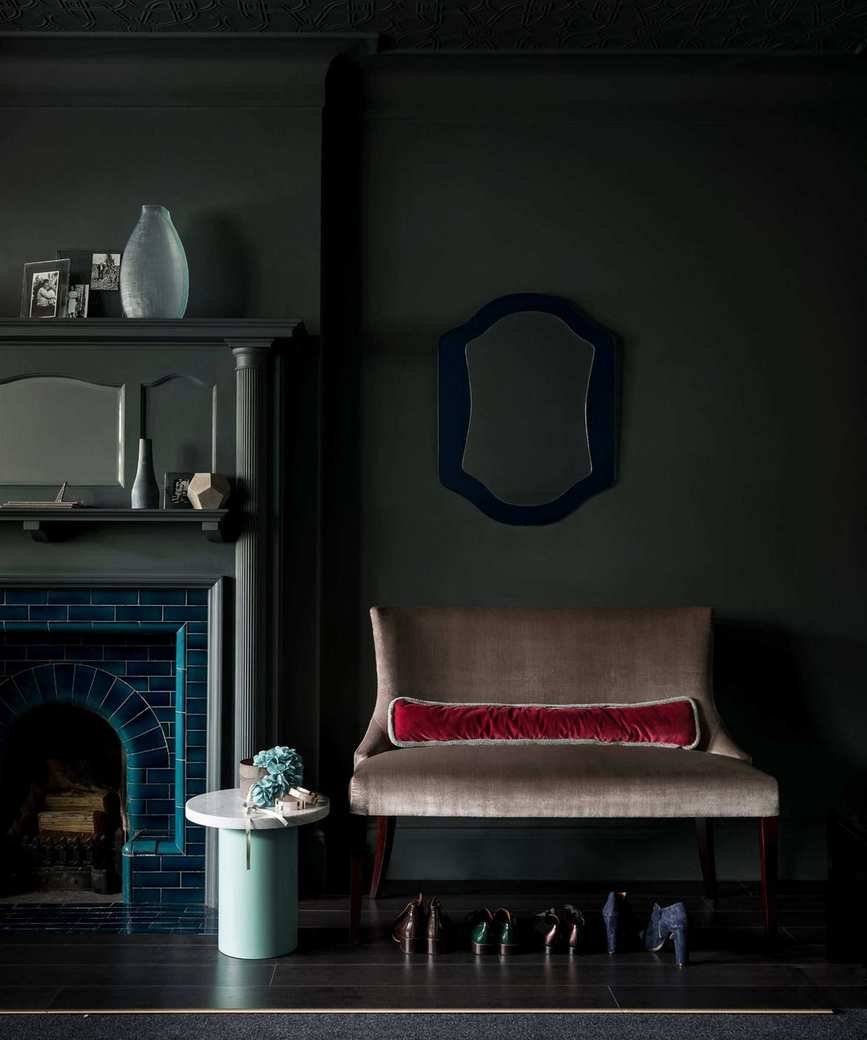 A dark living room with dark greeny grey walls, a blue fireplace with matching dark mantelpiece, a large beige upholstered chair with a row of shoes underneath, and a green circular table with white top to the left of it.