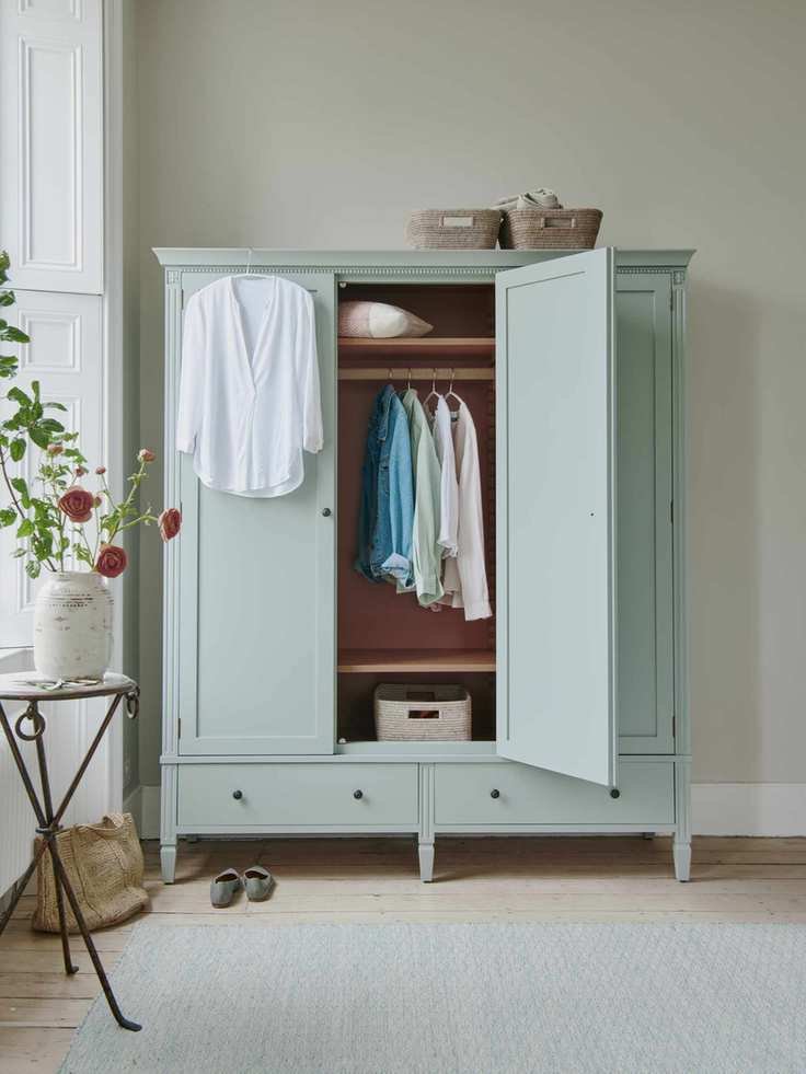 bedroom with aqua freestanding wardrobe, clothes hanging, baskets on top, side table near window with flowers, aqua rug
