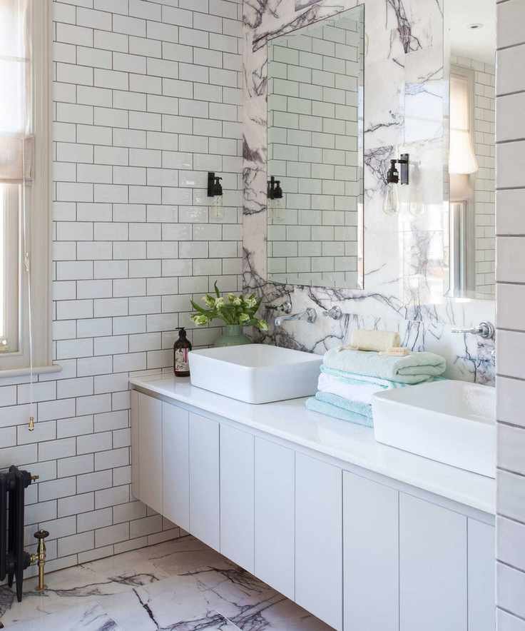 A white tiled bathroom with a long vanity in front of a marble wall