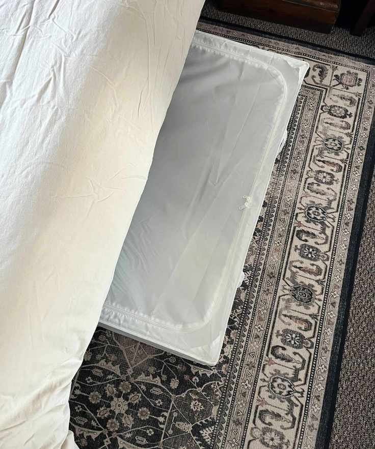 A white under-bed storage bag under a cream bed, on top of a patterned brown rug.
