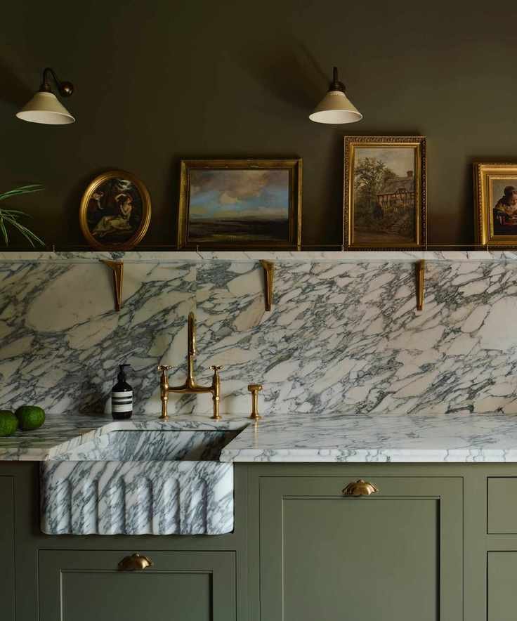 green and marble kitchen with sage green cabinetry, marble countertop, backsplash and shelf, green walls, marble sink, brass hardware, artwork on shelf, wall lights