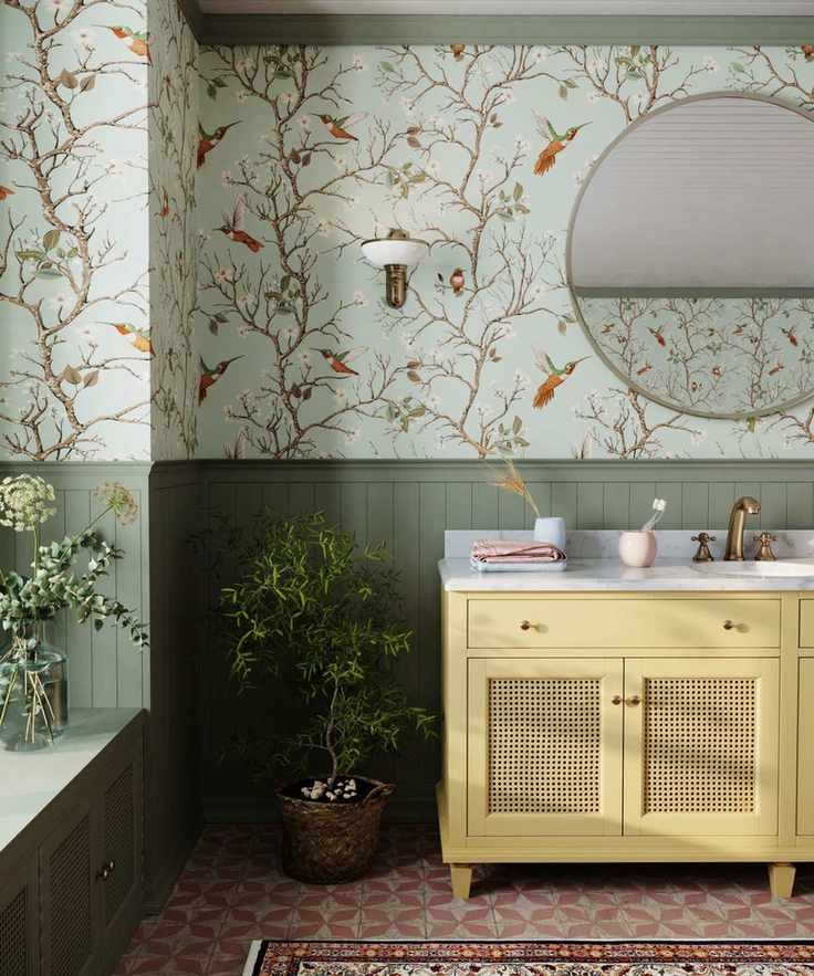Sage green half paneled bathroom with butter yellow vanity and the Bronte tree and bird wallpaper. There are potted plants, a window seat, and tiled geometric orange floor tiles with a bathroom rug