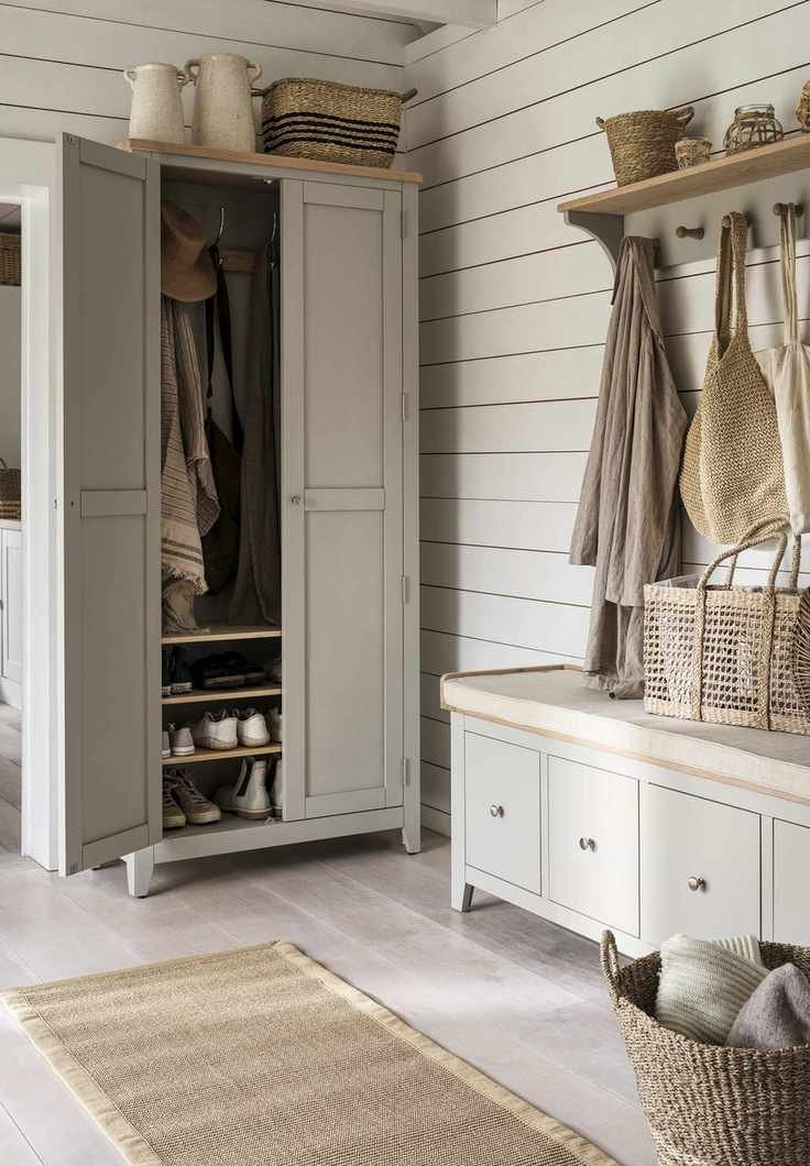 A gray painted free-standing closet in a white ship-lap entryway, beside a matching shoe bench with drawer storage, and a wall rack with coats hanging