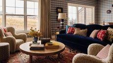 an open plan living area in a home in the Catskills with blue gingham wallpaper, a navy velvet couch, vintage red rug, sheepskin armchairs and a round wooden coffee table