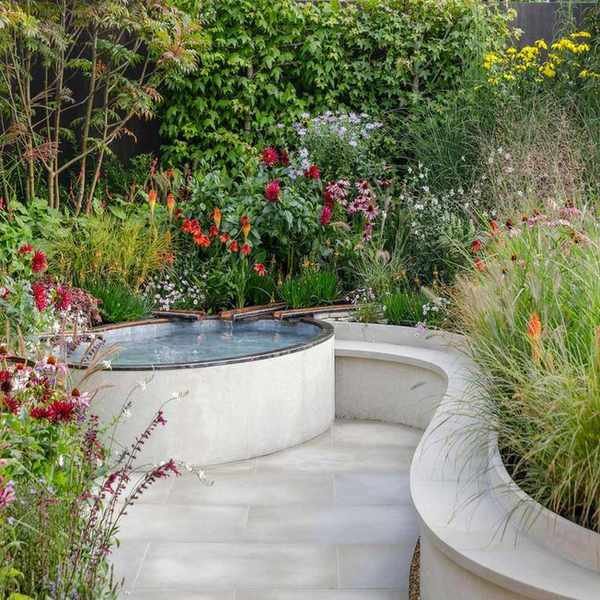 curved garden design with wall, paving and water feature, plus naturalistic planting with a hedge in the background