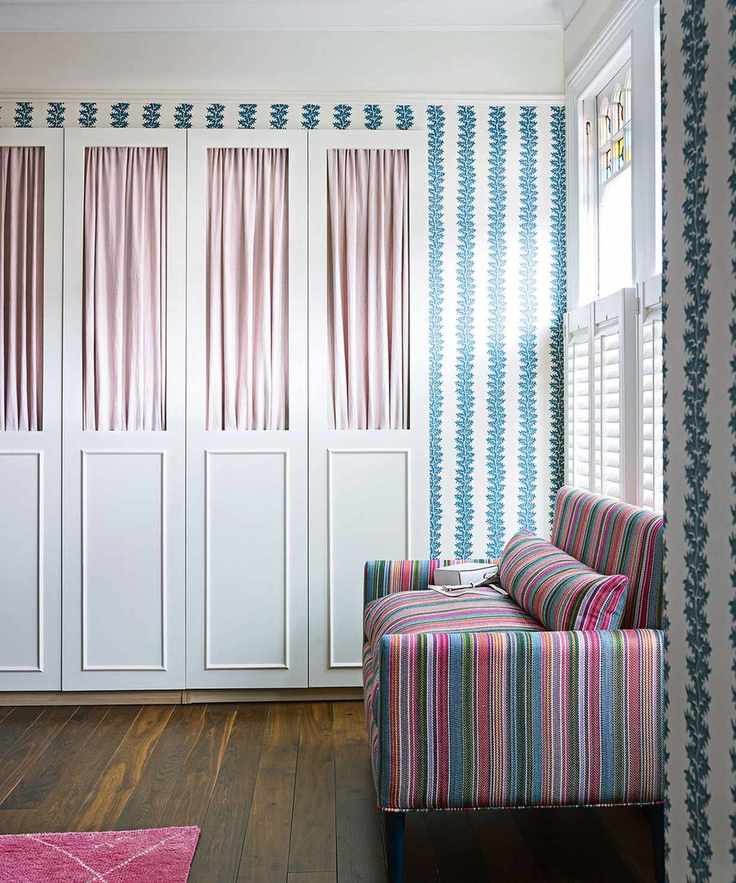 Bedroom with dark wooden floor colourful striped sofa, blue and white wallpaper and built in wardrobes, pink fabric curtains