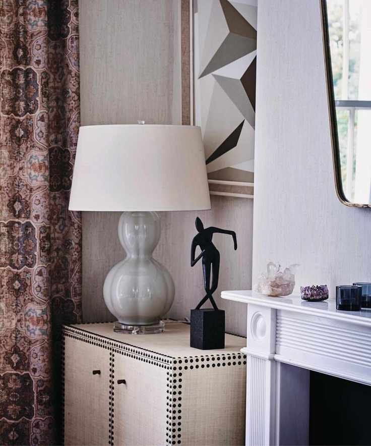 A living room with a grey lamp and shade on a small cupboard in an alcove beside a fireplace, and long patterned curtains.