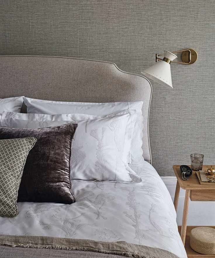 A neutral bedroom space with taupe walls, a grey brown bed with white bedding, assorted cushions, and a wooden bedside table. Behind are white painted baseboards, and the flooring is hardwood.