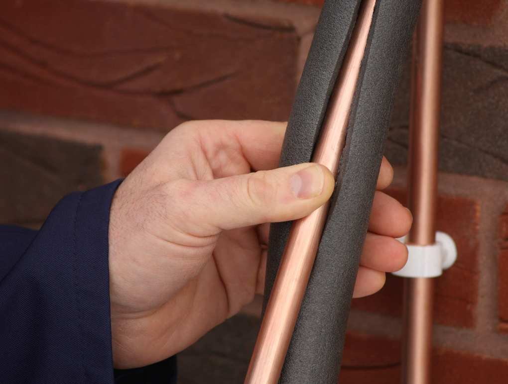 A man wrapping a copper pipe in a coat of foam insulation against a brick wall