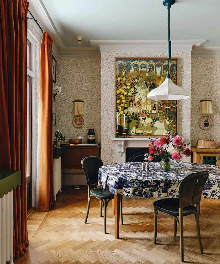 scandi maximalist style dining room with wallpapered walls, a large fireplace with folk art hung above, dark orange velvet curtains, a dining table with blue and white printed tablecloth and a white fabric pendant light