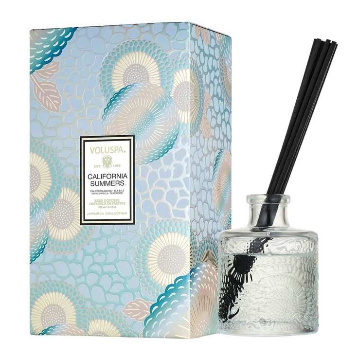 A small glass reed diffuser with black reeds next to a blue patterned packaging box