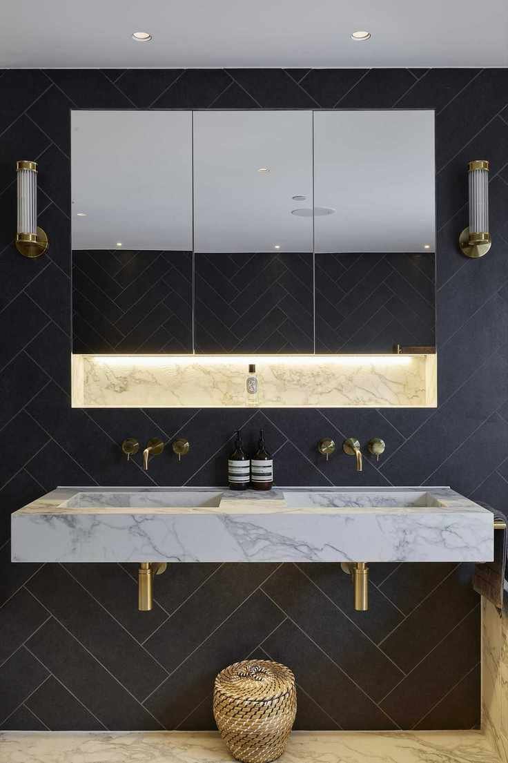 An example of dark bathroom ideas showing a marble basin with gold taps, gold wall lights and a mirror, and navy herringbone tiles