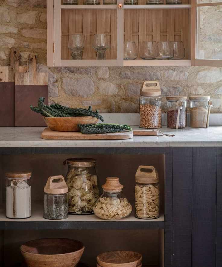 A rustic kitchen with white countertops, glass jars of dried food goods on the counter and shelves beneath, and dark blue painted cabinets.