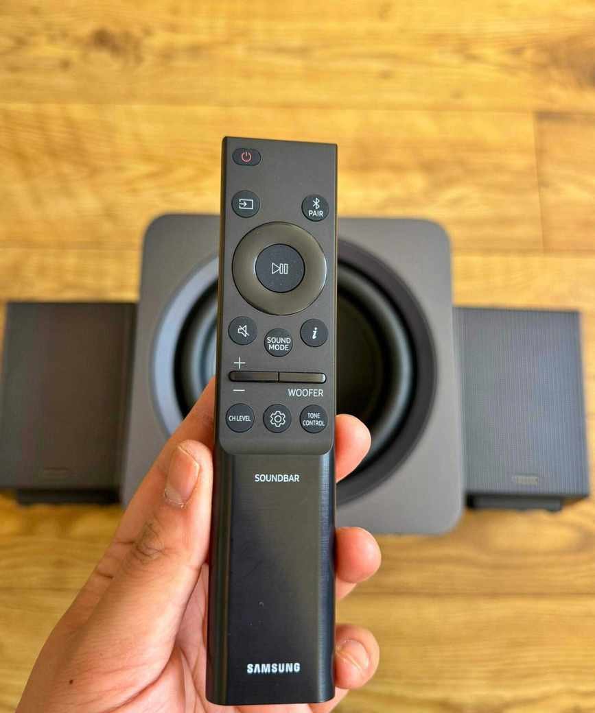Hand holding a soundbar remote above a subwoofer and two rear speakers on wooden floor.
