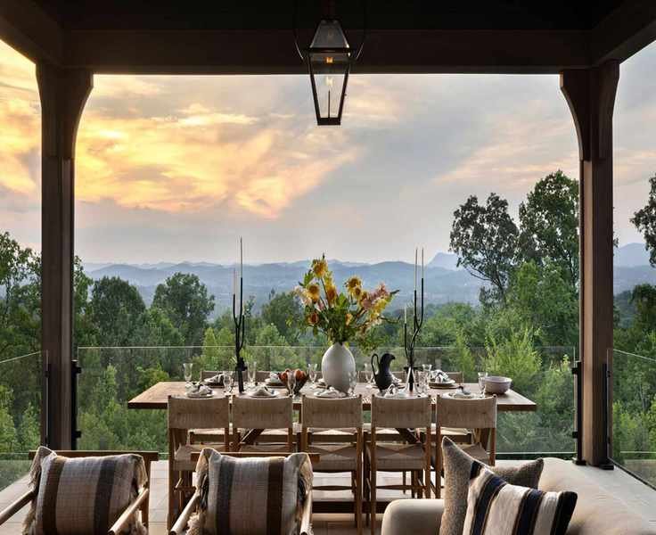 Covered outdoor dining space with wooden dining table and cream and wood chairs. The view is over the forest and the sun in setting