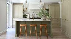 A kitchen with sage green cabinets and a darker green island. A large stone vase with green steps on the island beside a sink, and two glass pendants hanging from the ceiling above.